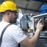 Factory worker in yellow hard hat operating industrial control panel for AI predictive maintenance system